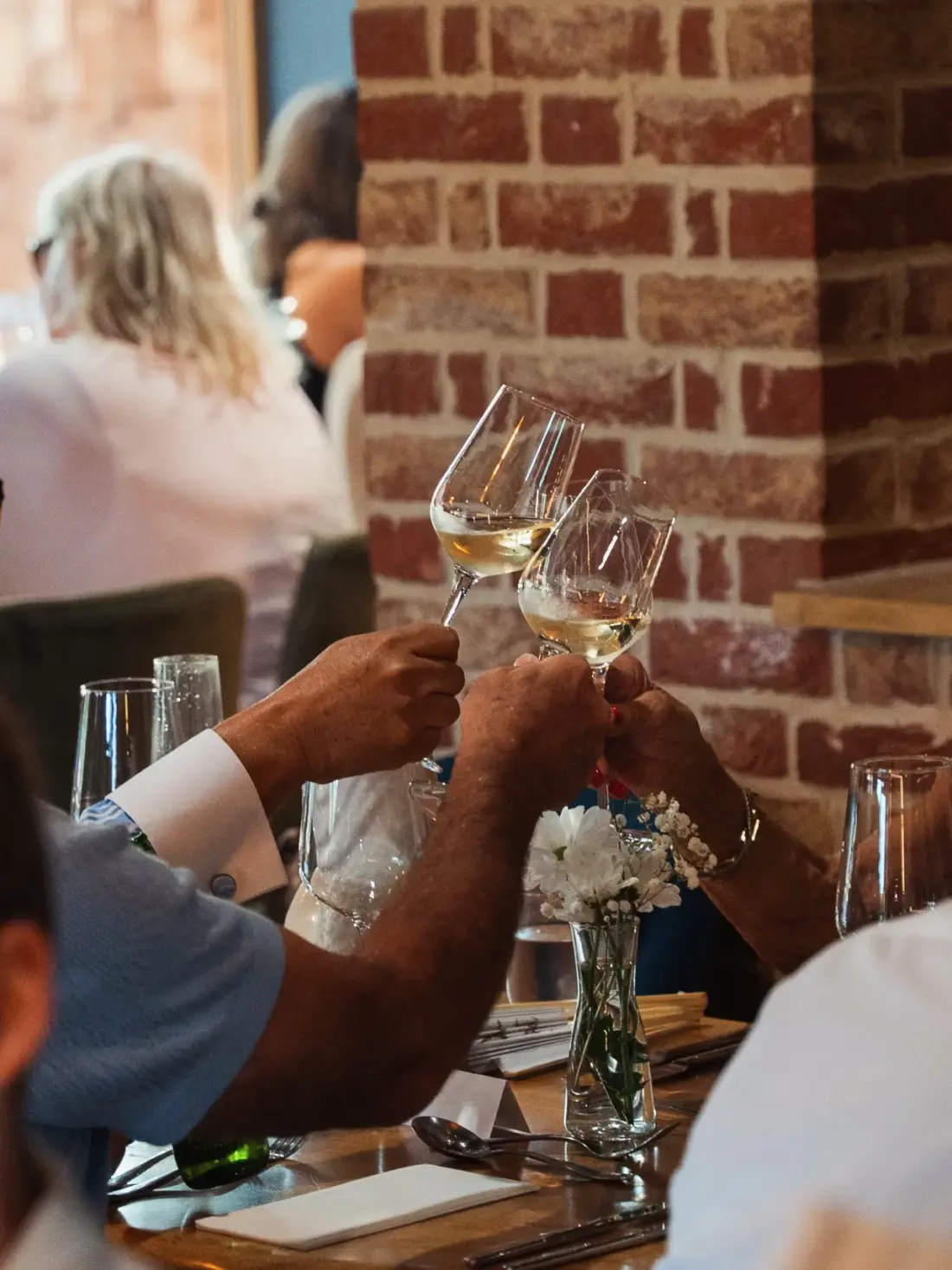 Two hands clinking glasses of white wine in front of a brick wall